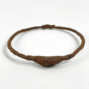 May include: A circular, rusted iron torque necklace featuring a twisted metal band design. The piece has a prominent, thickened central section and a simple knotted closure mechanism at the top, displaying a weathered, oxidized brown texture throughout.