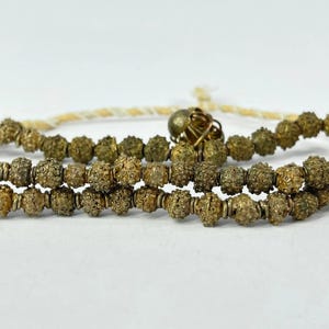 May include: Close-up of a necklace featuring intricately detailed, textured, gold-colored beads. The beads are strung together with a cream-colored cord and a gold-colored clasp. The necklace is displayed against a white background.