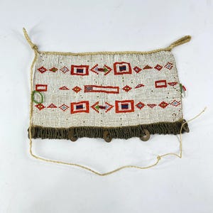 May include: A beaded bag with a cream-colored background and geometric patterns in red, blue, and green. The bag has a dark brown fringe at the bottom and is adorned with wooden buttons. It has a woven cord for hanging.