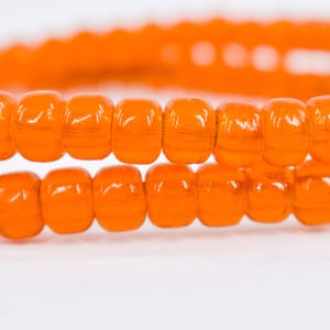 Könnte beinhalten: Nahaufnahme von zwei Strängen orangefarbener Perlen. Die Perlen sind fassförmig und haben eine glänzende Oberfläche. Der Hintergrund ist weiß und hebt die leuchtende Farbe der Perlen hervor. Die Perlen sind aufgefädelt, was darauf hindeutet, dass sie für die Schmuckherstellung bestimmt sind.