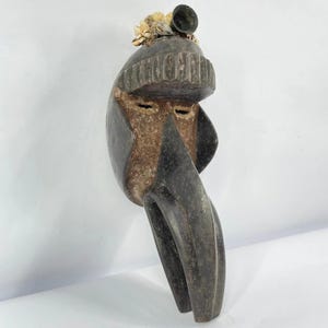 May include: A carved wooden mask with a dark gray finish. The mask features a prominent beak-like nose, small eye slits, and a hat-like structure on top, adorned with small, light-colored embellishments and a dark, round object.