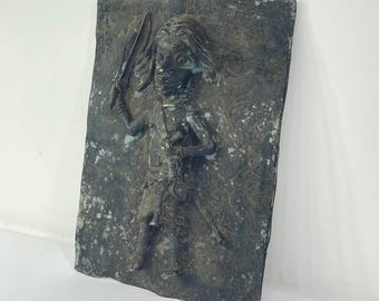 May include: A bronze-toned relief sculpture depicting a figure holding a sword and a smaller object. The figure has long hair and is wearing what appears to be armor. The rectangular plaque has a textured surface and a weathered appearance.