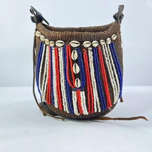 May include: A woven brown basket bag with a decorative beaded design. The bag features vertical strands of red, white, and blue beads, accented with seashells. Leather straps and accents complete the design.