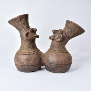 Puede incluir: Una escultura de cerámica marrón que representa dos figuras unidas en la base. Cada figura tiene un cuerpo bulboso, un cuello texturizado y una cabeza con un pico prominente y un sombrero alto y texturizado. La escultura tiene un aspecto rústico y artesanal.