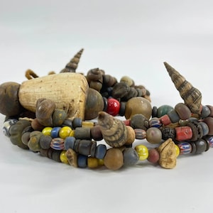 May include: A multi-strand necklace featuring a collection of assorted beads in various shapes, sizes, and colors, including brown, yellow, blue, red, and grey. The piece incorporates natural elements like small spiral seashells and carved bone.