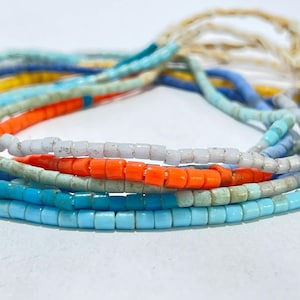 May include: A collection of multi-strand beaded necklaces featuring small cylindrical beads in vibrant shades of bright orange, sky blue, turquoise, white, and yellow. The strands are arranged in a layered formation, showcasing a variety of colors.