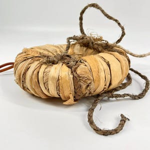 Puede incluir: Un objeto circular en forma de toroide construido con fibras vegetales secas de color tostado claro envueltas firmemente. La estructura está asegurada con una cuerda trenzada de color marrón oscuro que rodea el perímetro y termina en extremos sueltos.