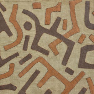 May include: Close-up of a textile with a repeating pattern of abstract shapes in shades of brown and orange against a beige background. The shapes appear to be hand-stitched onto the fabric, creating a textured effect. The design has a tribal or ethnic aesthetic.