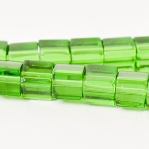 May include: Close-up of a strand of bright green, rectangular glass beads. The beads are translucent and have a slightly rounded shape. The beads are strung together, creating a line of vibrant color.