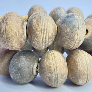 May include: A collection of spherical, weathered clay beads arranged in rows. Each bead features a matte, earthy brown and grey finish with subtle, incised parallel line details encircling the center. The beads are strung together on a light-colored cord.