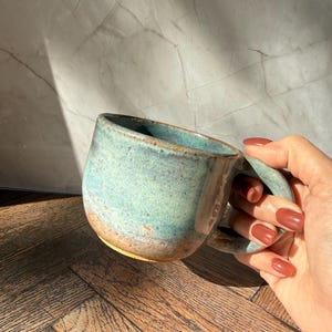 May include: A handmade ceramic mug with a rounded shape and a large handle. The mug features a turquoise glaze with brown and tan accents. The mug is designed for hot beverages and is suitable for home use.