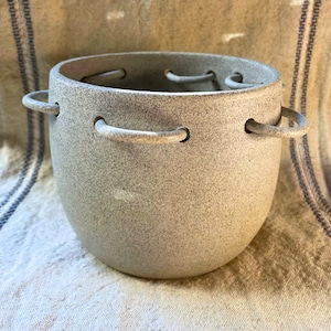May include: A speckled, light gray ceramic pot with two curved handles on the sides. The pot has a rounded shape and a wide opening. There are small holes around the rim. The pot is sitting on a textured, off-white surface.