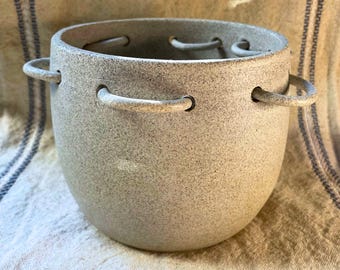 May include: A speckled, light gray ceramic pot with two curved handles on the sides. The pot has a rounded shape and a wide opening. There are small holes around the rim. The pot is sitting on a textured, off-white surface.