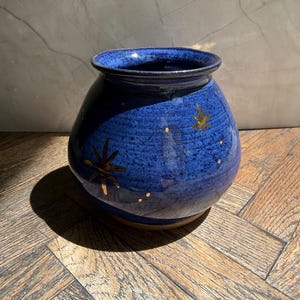 May include: A round, blue ceramic vase with a wide opening. The vase is decorated with gold star designs and small gold dots. The vase sits on a wooden surface and has a dark shadow.