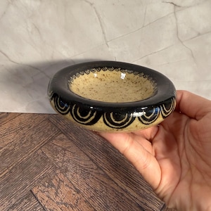 May include: A small, round ceramic bowl with a speckled beige interior and a black rim. The rim is decorated with a repeating pattern of black semi-circles. The bowl is held in a hand.