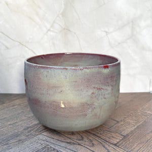 May include: A round ceramic bowl featuring a smooth, glazed finish with a blended color palette of muted sage green, soft lavender, and deep burgundy tones. The interior rim shows a darker red glaze, while the exterior displays a mottled texture.