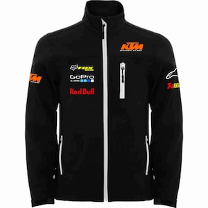 Puede incluir: Chaqueta negra con cremallera, cremalleras blancas y cuello alto. La chaqueta presenta el logotipo de KTM Racing Team en naranja, junto con los logotipos de Fox, GoPro y Red Bull. Logotipos adicionales en las mangas.