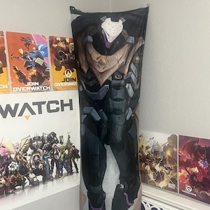 May include: A rectangular body pillow with a detailed illustration of a character from the video game Overwatch. The character is in a dark, armored suit with purple and white details. The pillow is propped against a wall.