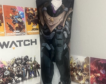 Op de afbeelding: Een rechthoekig bodykussen met een gedetailleerde illustratie van een personage uit de videogame Overwatch. Het personage draagt een donker, gepantserd pak met paarse en witte accenten. Het kussen leunt tegen een muur.