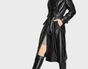 Op de afbeelding: Een lange, zwarte leren trenchcoat met een ceintuur in de taille en een kraag van imitatiebont. De jas heeft een dubbelborstig ontwerp en wordt gecombineerd met zwarte leren kniehoge laarzen. Het model draagt een rok onder de jas.