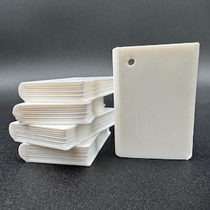 May include: A stack of four small, white, rectangular book-shaped objects with rounded spines, positioned next to a single, upright, white, rectangular tag featuring a circular hole at the top corner, all with a smooth, matte finish.