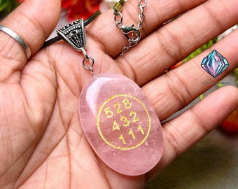 Puede incluir: Un colgante de cuarzo rosa con un diseño circular dorado que contiene los números 528, 432 y 111. El colgante, de forma ovalada, cuelga de un cordón negro y una cadena plateada.