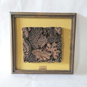 May include: A square, dark-toned metal batik press featuring an intricate peacock and floral pattern, mounted on a mustard yellow fabric background within a dark wooden frame. A small gold-colored plaque at the bottom reads BATIK PRESS.