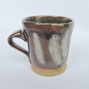 Può includere: Tazza in ceramica fatta a mano con smalto marrone e bianco. La tazza ha una base arrotondata e un manico curvo. L'esterno presenta un motivo unico a spirale di marrone e bianco. Il bordo è marrone scuro e la base è color cuoio chiaro.