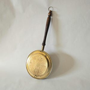 Puede incluir: Calientacamas antiguo de latón con un mango de madera oscura y un pequeño anillo de metal. El calentador de latón tiene un diseño ornamentado y grabado en la superficie de calentamiento circular. Un artículo de decoración vintage.