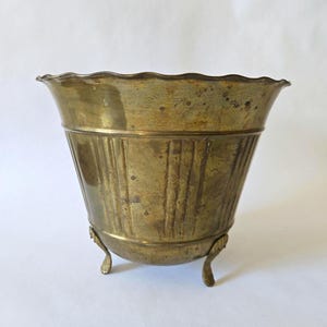 May include: A vintage brass planter with a scalloped rim and vertical ribbed detailing. The planter rests on three curved feet. The aged brass has a warm, golden hue, suggesting it is an antique or vintage piece.