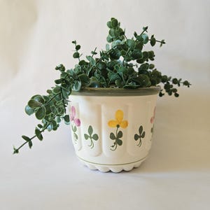 May include: A cream-coloured ceramic plant pot with a scalloped edge, decorated with painted flowers in pink and yellow, and green shamrock designs. The pot contains a cascading green plant. The rim of the pot is green.