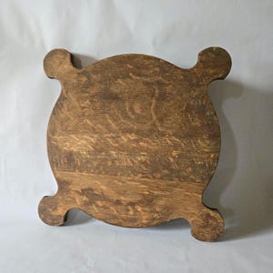 May include: A round, wooden trivet with a natural wood grain finish. The trivet has four curved, decorative feet. The wood has a warm, brown tone, and the trivet appears to be vintage or antique.