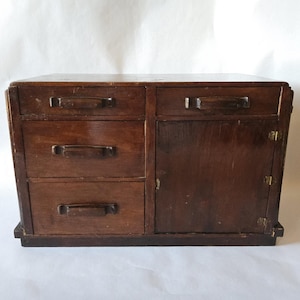 May include: A small, vintage wooden cabinet with a dark brown finish. It features three drawers on the left, a single door on the right, and a rectangular shape. The cabinet has small, dark handles and hinges.