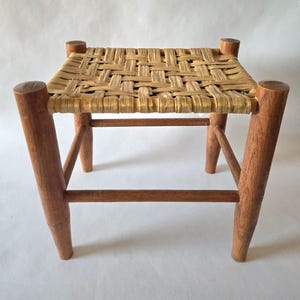 May include: A small, square wooden stool with a woven seat. The seat is made of light brown woven material. The legs are made of solid wood and are a medium brown color. The stool is a simple, rustic design.
