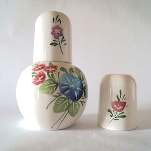 May include: A white ceramic water pitcher and cup set. The pitcher has a bulbous base and a lid, both decorated with hand-painted flowers in shades of red, blue, and purple. The cup has a similar floral design.