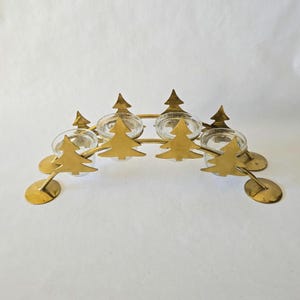 May include: A gold-colored Christmas tree candle holder with six clear glass votive holders. The candle holder features stylized Christmas tree cutouts and circular base supports. The design is symmetrical and festive, perfect for holiday decor.