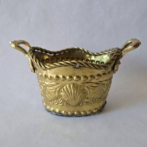 May include: A small, gold-colored metal basket with two curved handles. The basket's exterior features embossed seashell and floral designs, with a rope-like trim around the rim and a row of small, round embellishments. The basket is approximately 4 inches wide.