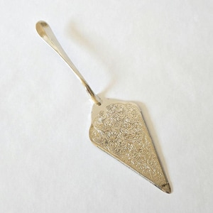 May include: Silver cake server with a decorative handle and a triangular blade. The blade features an ornate, embossed floral pattern. The server has a serrated edge for cutting and serving cake slices. The server is made of a shiny, reflective metal.