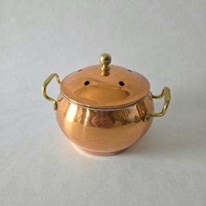 Peut inclure: Un pot de couleur cuivre avec un couvercle et deux poignées en laiton. Le couvercle comporte un petit bouton en laiton et plusieurs petits trous. Le pot est rond avec une finition brillante.