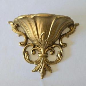 Könnte beinhalten: Eine goldfarbene Wandleuchte mit einer muschelartigen Schale und aufwendigen, sich windenden Blatt- und Blumendetails. Die Leuchte hat ein Fleur-de-Lis-Design am Boden und besteht aus einem metallischen Material. Das Design ist klassisch und dekorativ.