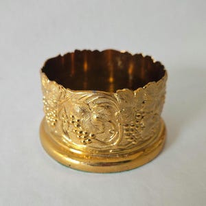 May include: A gold-colored metal wine coaster with a detailed embossed grape and leaf design. The coaster has a round base and a scalloped rim. The design is repeated around the entire circumference of the coaster.