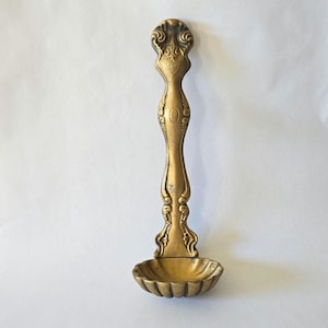 May include: A vintage brass spoon with an ornate handle and a shell-shaped bowl. The handle features decorative scrollwork and a crest. The spoon is a warm gold color and appears to be made of metal. The spoon is likely used for serving or display.