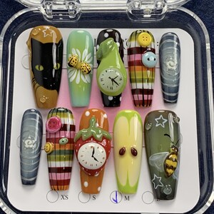 May include: A set of ten press-on nails featuring diverse designs including a black cat, a ladybug on a daisy, a pear-shaped clock, plaid patterns with buttons, a strawberry clock, a sliced apple, and a bee with white stars on a dark background.