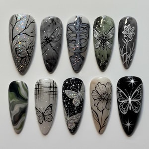 May include: A set of ten almond-shaped press-on nails featuring various designs including black line art of butterflies, dragonflies, flowers, and a rib cage. The color palette includes shades of black, gray, white, and olive green with glitter accents.