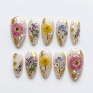 May include: A set of ten press-on nails with a milky white base, decorated with pressed flowers including pink roses, yellow buttercups, blue forget-me-nots, and white daisies, accented with gold leaf flakes, white swirling lines, and small pearls.