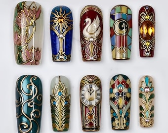 May include: A set of ten press-on nails featuring intricate designs including a pink lotus, a sun, a white swan, a crescent moon, a clock face, peacock feathers, and ornate gold filigree patterns with embedded rhinestones and colorful enamel.