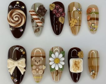 May include: A set of ten almond-shaped press-on nails featuring a brown and beige color palette. Designs include 3D embellishments like a chocolate swirl, candy cane, teddy bear, daisy, marshmallow, bow, and rhinestones over plaid patterns.