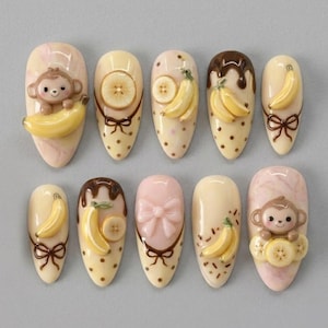May include: A set of ten press-on nails featuring a cream and pink marble base with 3D decorations including brown monkeys, yellow bananas, sliced banana rounds, brown chocolate drips, brown bows, and a pink bow, all arranged in two horizontal rows.