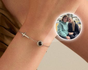 May include: A silver bracelet featuring a small airplane charm and a black gemstone. The bracelet is worn on a wrist, with a small circular photo of two people in the background.