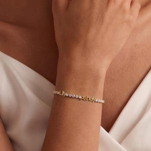 Könnte beinhalten: Ein zartes goldenes Armband mit den Namen "Lily" und "Ashey" in kleinen, eleganten Buchstaben geschrieben. Das Armband ist mit kleinen, funkelnden Verzierungen versehen. Der Schmuck wird am Handgelenk getragen.